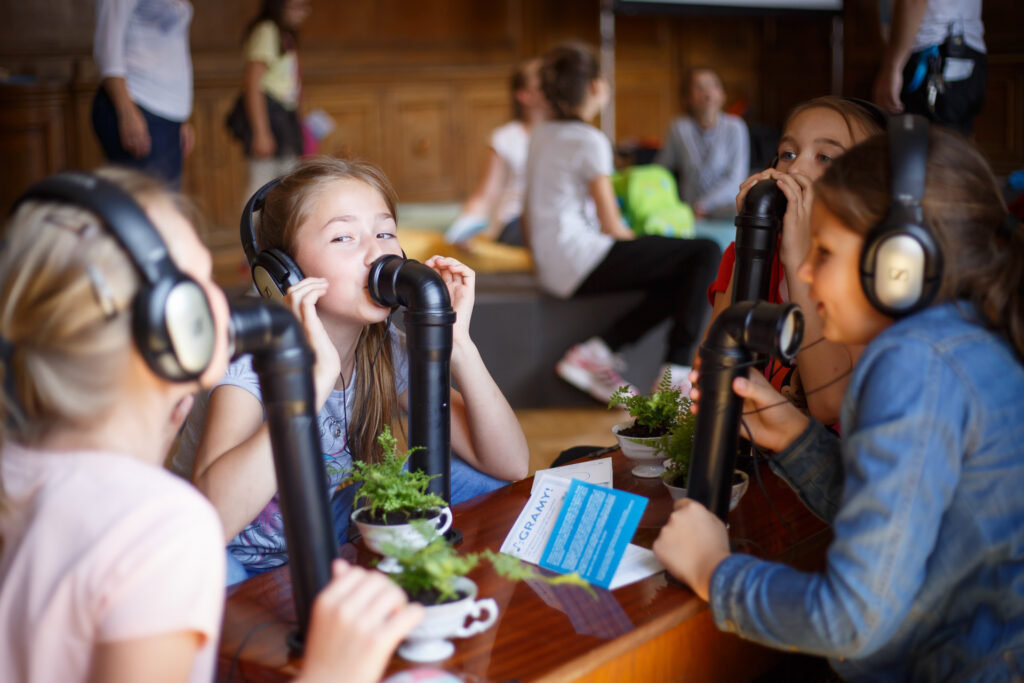 Na zdjęciu grupa dziewczynek przy blacie z przytwierdzonymi do niego czarnymi rurami. Dziewczynki mówią do otworów w rurach i każda z nich ma założone słuchawki.