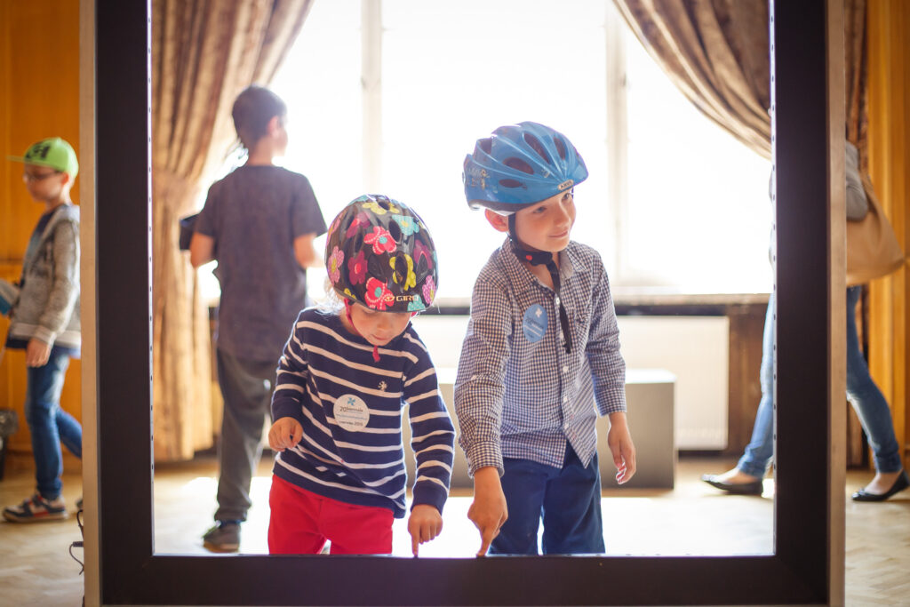 Na zdjęciu chłopiec i dziewczynka w kaskach rowerowych, Oboje stoją przed pustą ramą od obrazu i dotykają palcami jej brzegu.