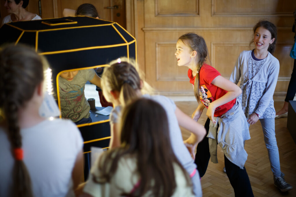 Na zdjęciu konstrukcja przypominająca stary telewizor z ekranem po czterech stronach. W miejscu ekranów są lusterka. Dzieci przeglądają się w nich i śmieją.