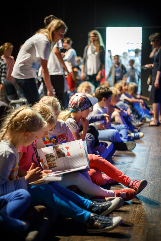 Na zdjęciu grupa dzieci na widowni. Dzieci siedzą na poduszkach na podłodze. Niektóre przeglądają książeczki.