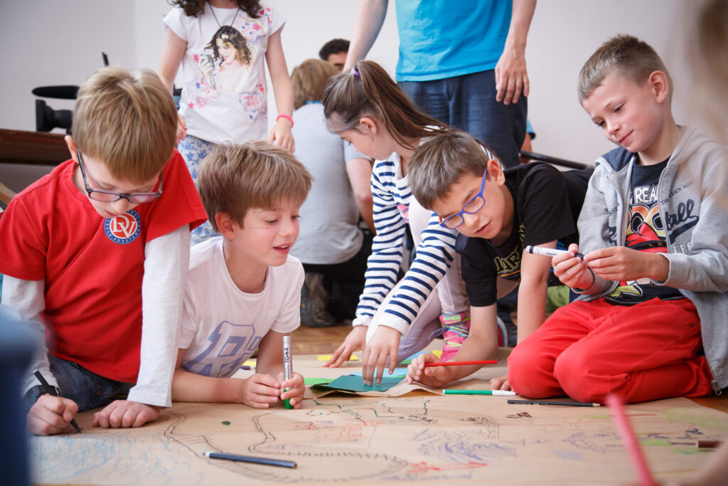 Na zdjęciu grupa dzieci siedzących na podłodze rysuje plakat.