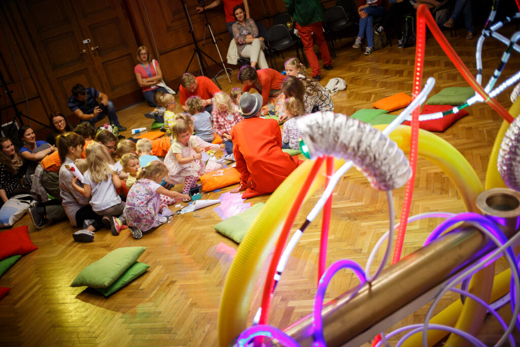 Grupa dzieci i dorosłych siedzi na podłodze. Niektórzy siedzą kolorowych poduszkach. Wspólnie wykonują prace plastyczne, wycinają.