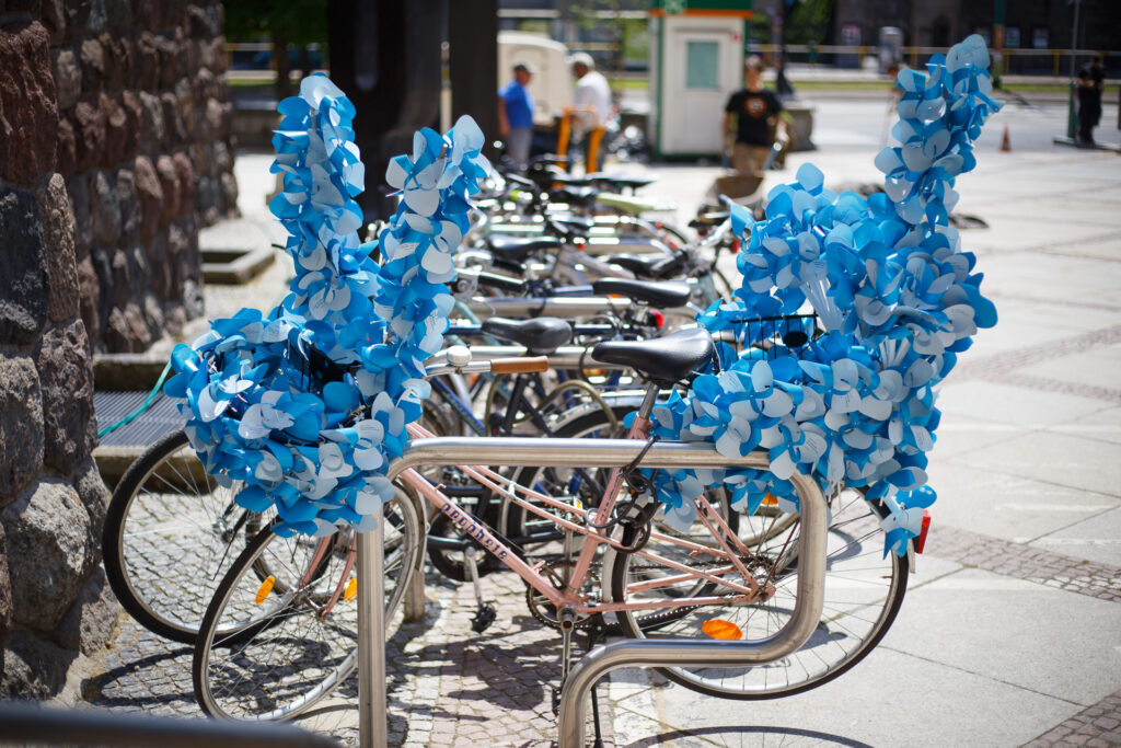 Na zdjęciu stojaki z rowerami na zewnątrz. Jeden z nich ma obficie ozdobiony przód i tył niebieskimi wiatraczkami
