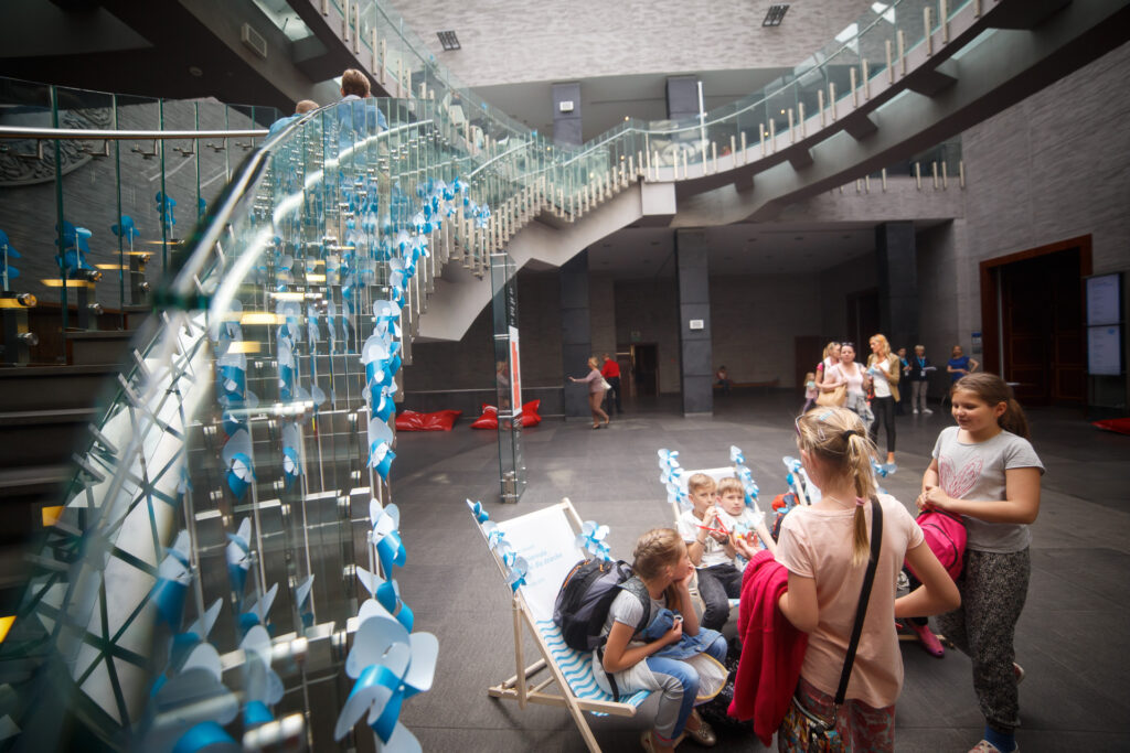 Na zdjęciu schody na piętro CK Zamek z przeszklonymi poręczami. Ozdobione są niebieskimi wiatraczkami Biennale. W holu leżaki Biennale, na których siedzą dzieci.