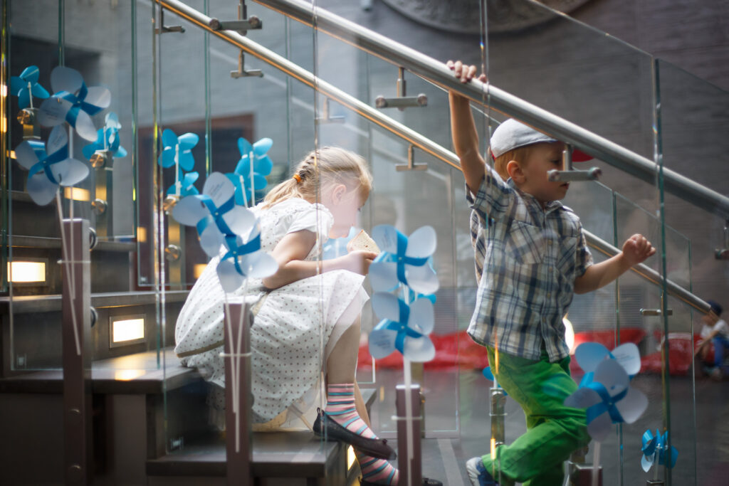 Na zdjęciu dwoje dzieci na schodach z przeszklonymi poręczami w CK Zamek ozdobionych niebieskimi wiatraczkami Biennale. Dziewczynka siedzi na schodach, a chłopiec schodzi na dół