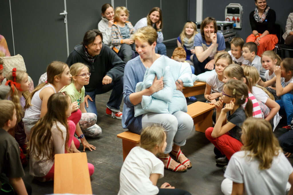 Na zdjęciu na ławce siedzi kobieta i trzyma dwie niebieskie poduszki, uśmiecha się szeroko. Wokół niej na takich samych ławkach siedzą dzieci i dorośli, którzy na nią patrzą również się uśmiechając.