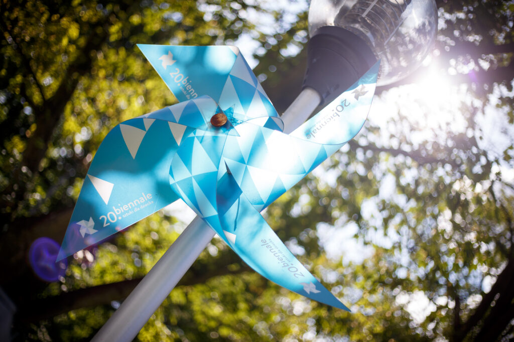 na zdjęciu wiatraczek 20. Biennale w kolorach niebieskiego. W tle drzewa.