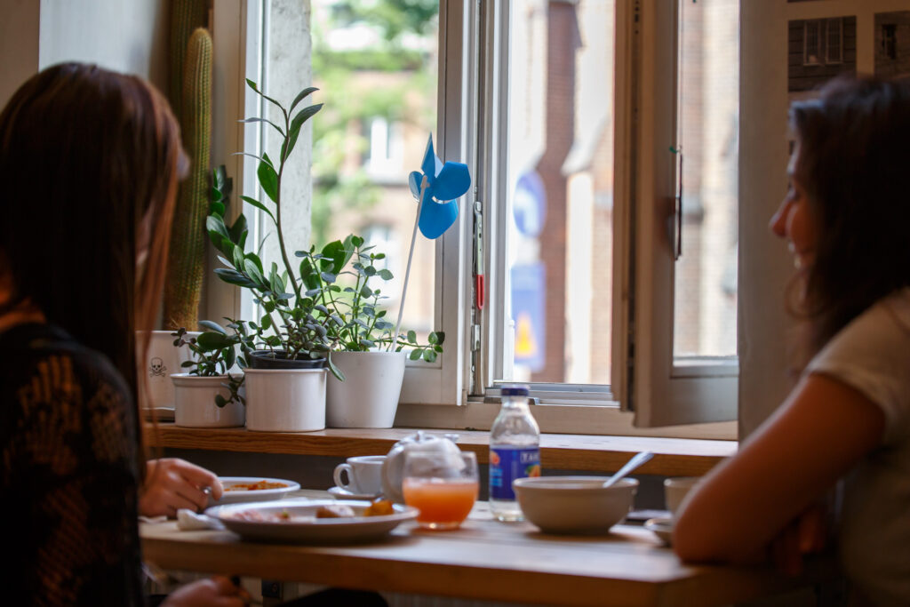 Na zdjęciu dwie kobiety odwrócone tyłem do obiektywu, siedzące przy stole. Przed stołem parapet i otwarte okno. Na parapecie kwiaty, a w jednej z doniczek niebieski wiatraczek Biennale.