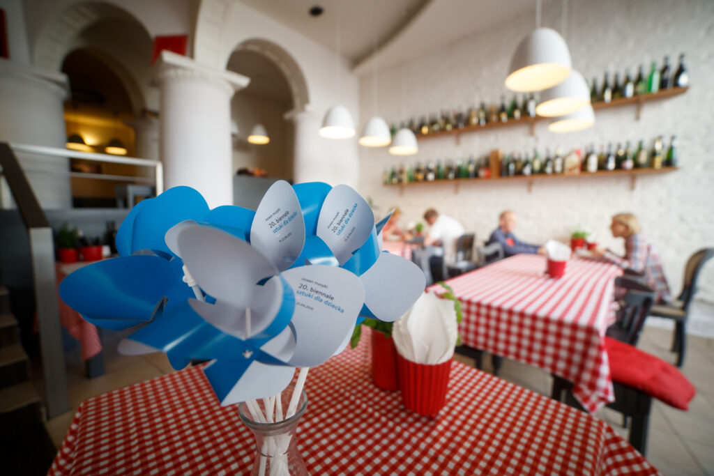 Na zdjęciu restauracja. Na jednym ze stołów wazon z niebieskimi wiatraczkami Biennale.