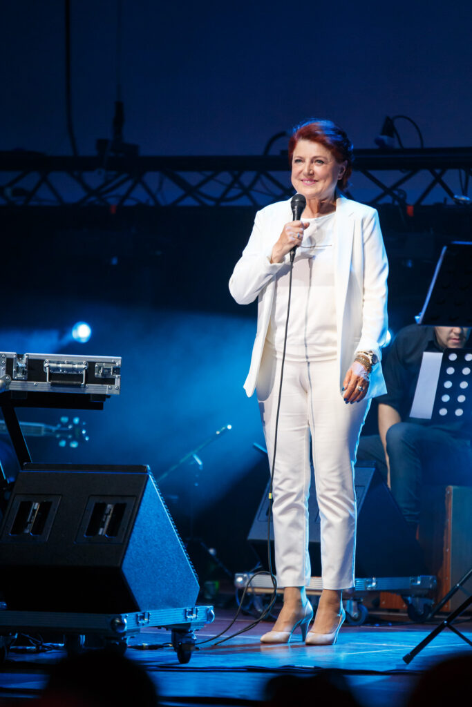 Na zdjęciu ubrana na biało kobieta w krótkich włosach stoi na scenie. Przy twarzy trzyma mikrofon i uśmiecha się szeroko