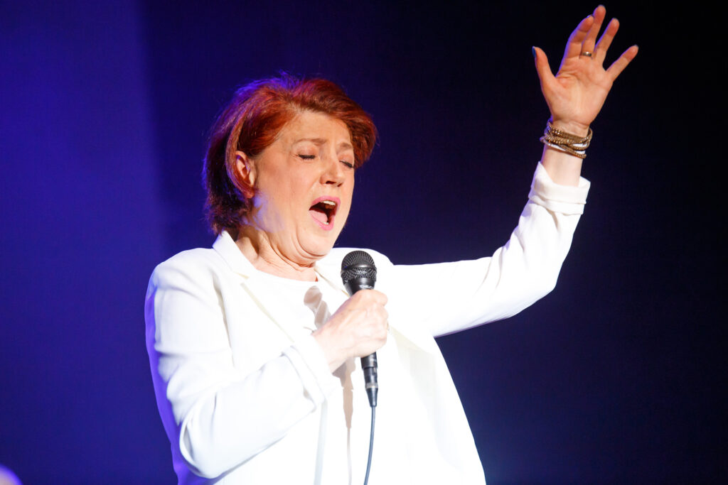 Na zdjęciu ubrana na biało konieta w krótkich włosach śpiewa do mikrofonu