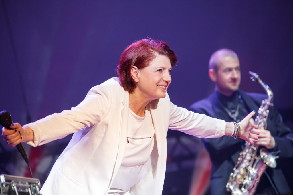 Na zdjęciu ubrana na biało kobieta w krótkich włosach. Stoi na scenie i kłania się po występie z uśmiechem. W tle zespół muzyczny.