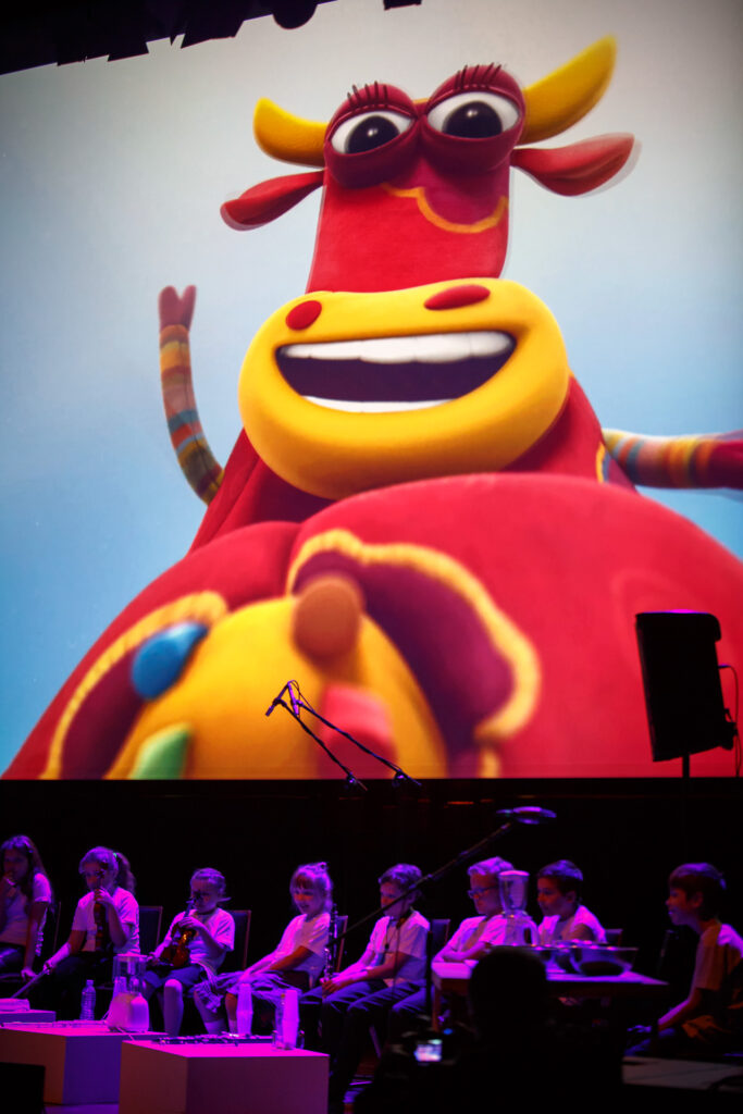 Na zdjęciu grupa dzieci siedząca w rzędzie na scenie. Za nimi duży ekran, na którym widać rysunkową krowę.