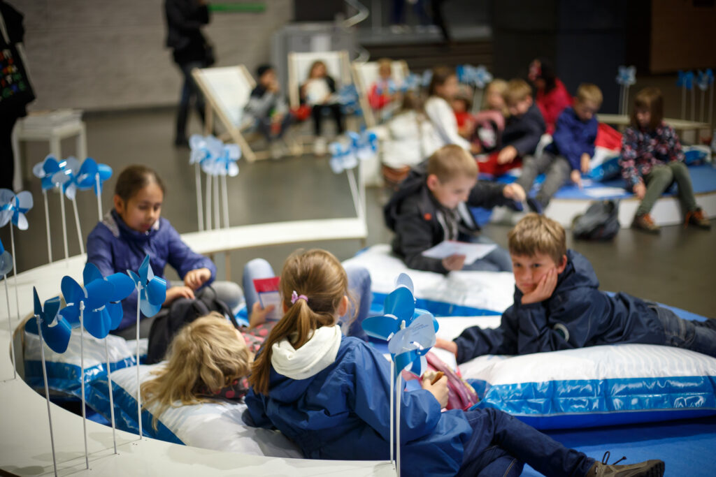 Na zdjęciu dzieci siedzące na niebieskich poduszkach w foyer CK Zamek. Przestrzeń ozdobiona jest niebieskimi wiatraczkami Biennale.