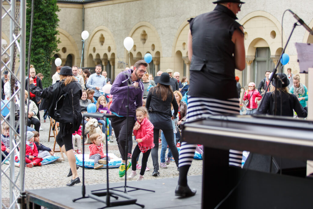 Dziedziniec CK Zamek. Dzieci i dorośli tańczą albo siedzą na poduszkach na ziemi. Dziedziniec ozdobiony jest niebieskimi i białymi balonami.