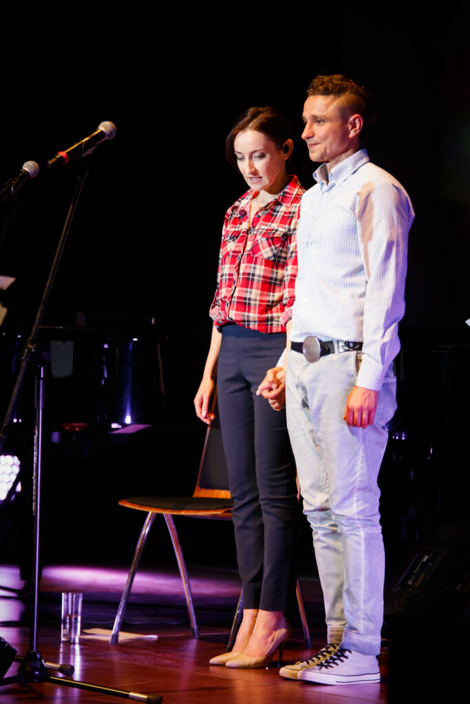 Na zdjęciu stojący obok siebie na scenie mężczyzna i kobieta, którzy trzymają się za ręce.