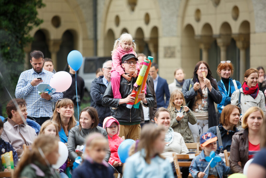Dziedziniec CK Zamek. Widownia dzieci i dorosłych. Na środku z tyłu mężczyzna stoi i trzyma dziewczynkę na barana. W dłoniach trzyma kolorowe tuby z papierowych rolek i bibuły. Mężczyzna po lewej stronie stoi i patrzy w telefon. W drugiej ręce trzyma biały balon. Kobieta po prawej stronie je lody i uśmiecha się.