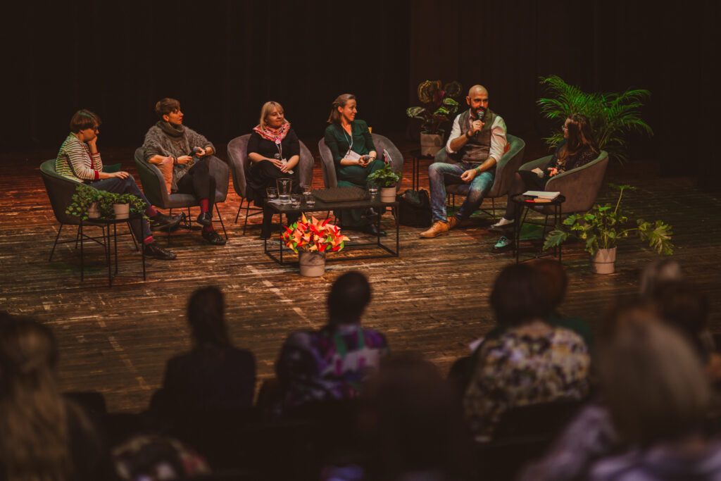 Na zdjęciu sześć osób siedzi na scenie i rozmawia. Słuchają ich ludzie na widowni.