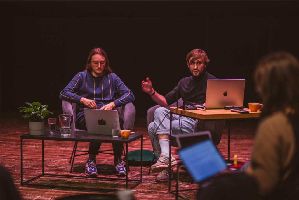 Na zdjęciu siedzi dwóch mężczyzn na scenie. Mają przed sobą laptopy.