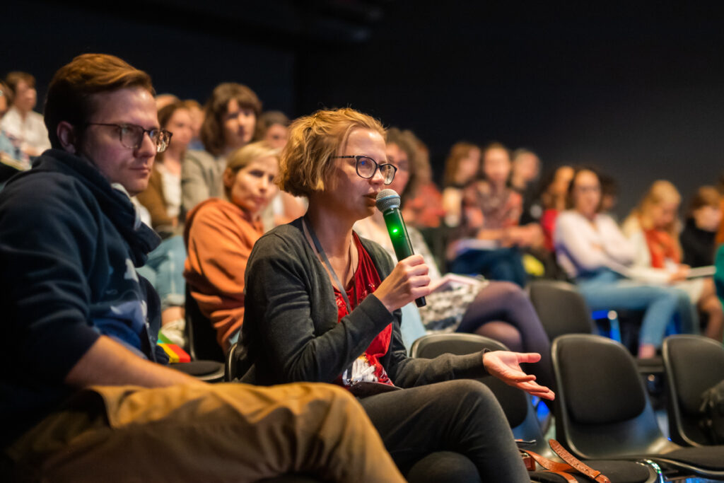 Panel. Na widowni siedzi grupa ludzi. Kobieta w krótkich włosach i okularach z czarnymi oprawkami mówi do mikrofonu.