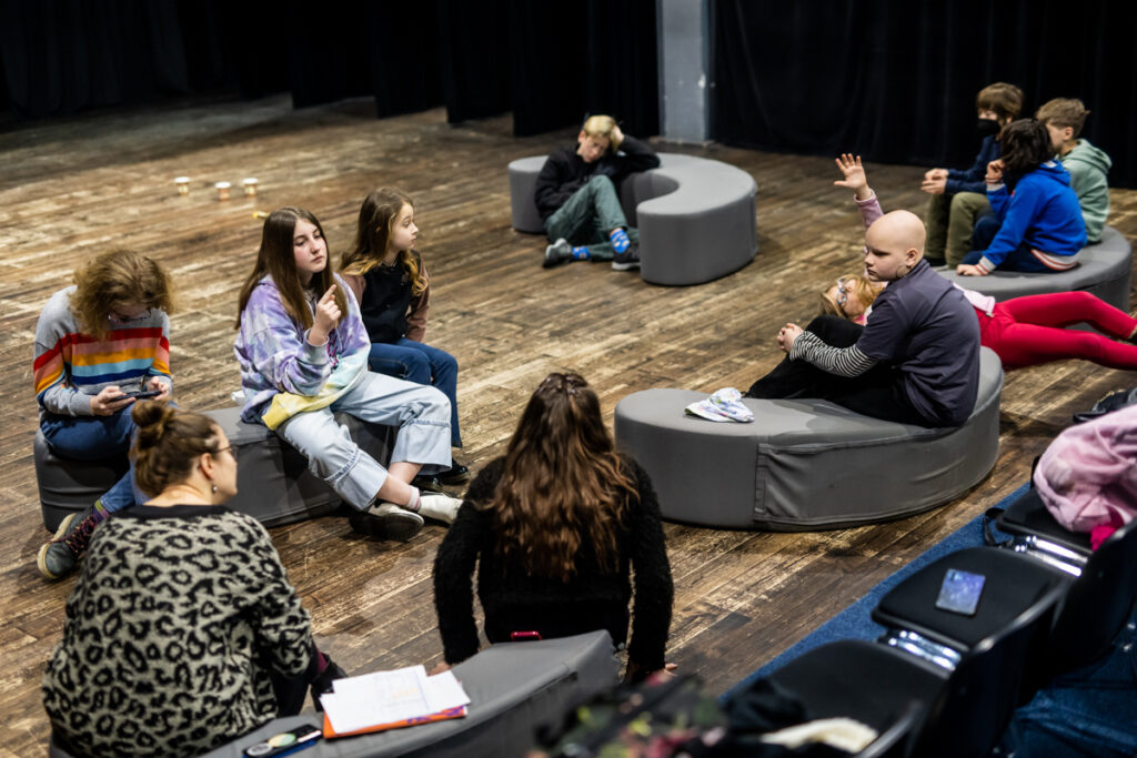 Osoby na scenie siedzą na poduszkach i rozmawiają.