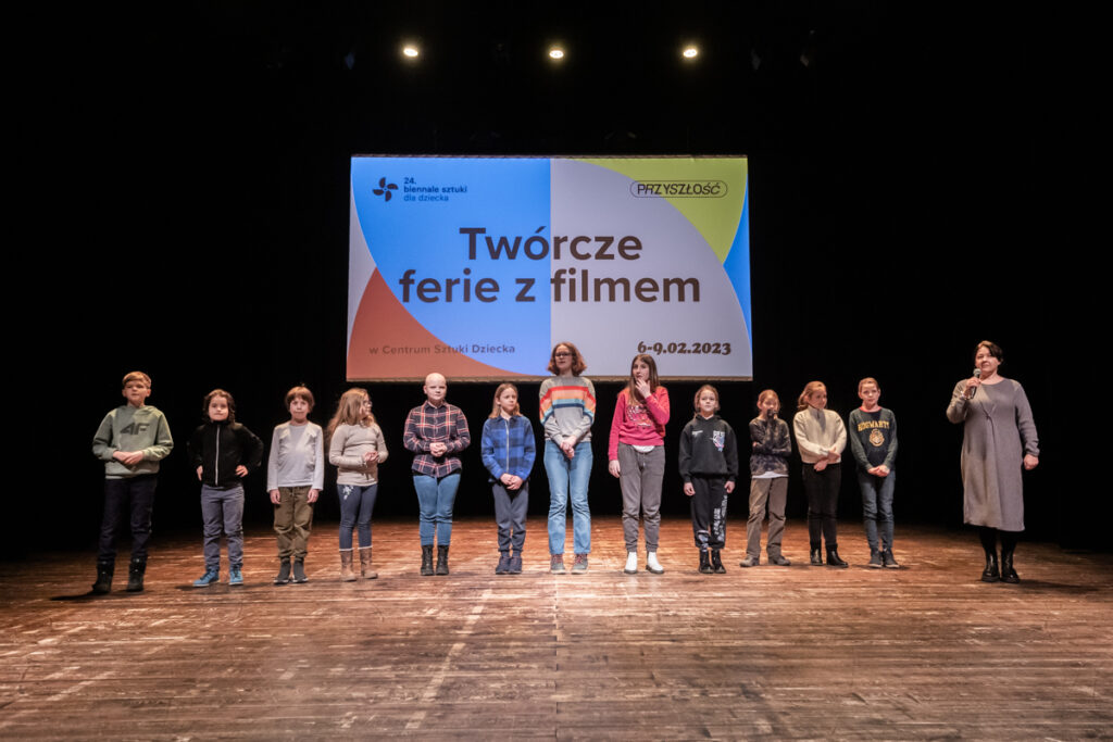 Osoby na scenie stoją przodem do widowni na tle kolorowej planszy wyświetlanej na ekranie.