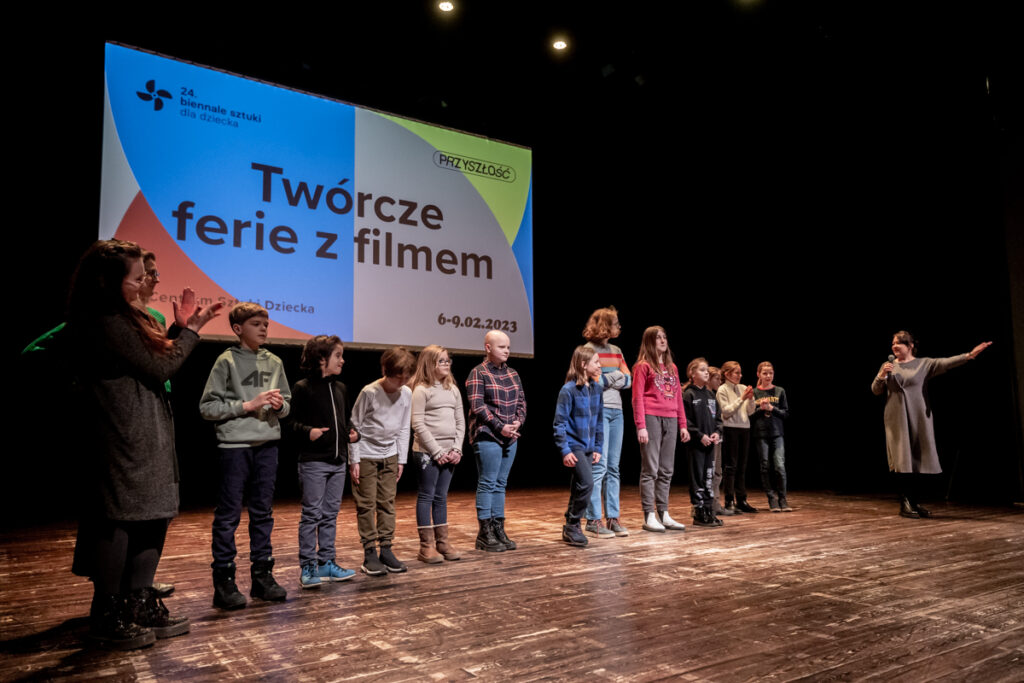 Osoby na scenie stoją przodem do widowni na tle kolorowej planszy wyświetlanej na ekranie.