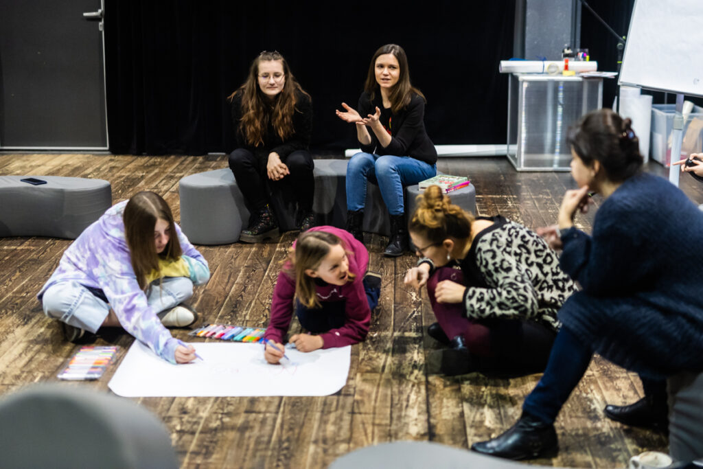 Osoby na scenie siedzą na poduszkach i rozmawiają.