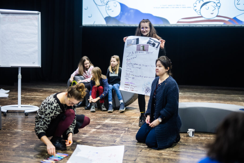 Osoby na scenie siedzą na poduszkach lub stoją i rozmawiają.