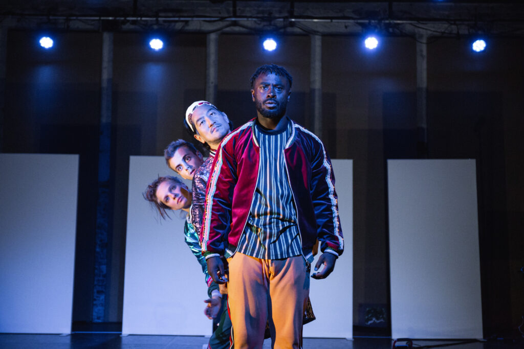 Czworo performerów na scenie stoi jedne za drugim i patrzy w stronę obiektywu.