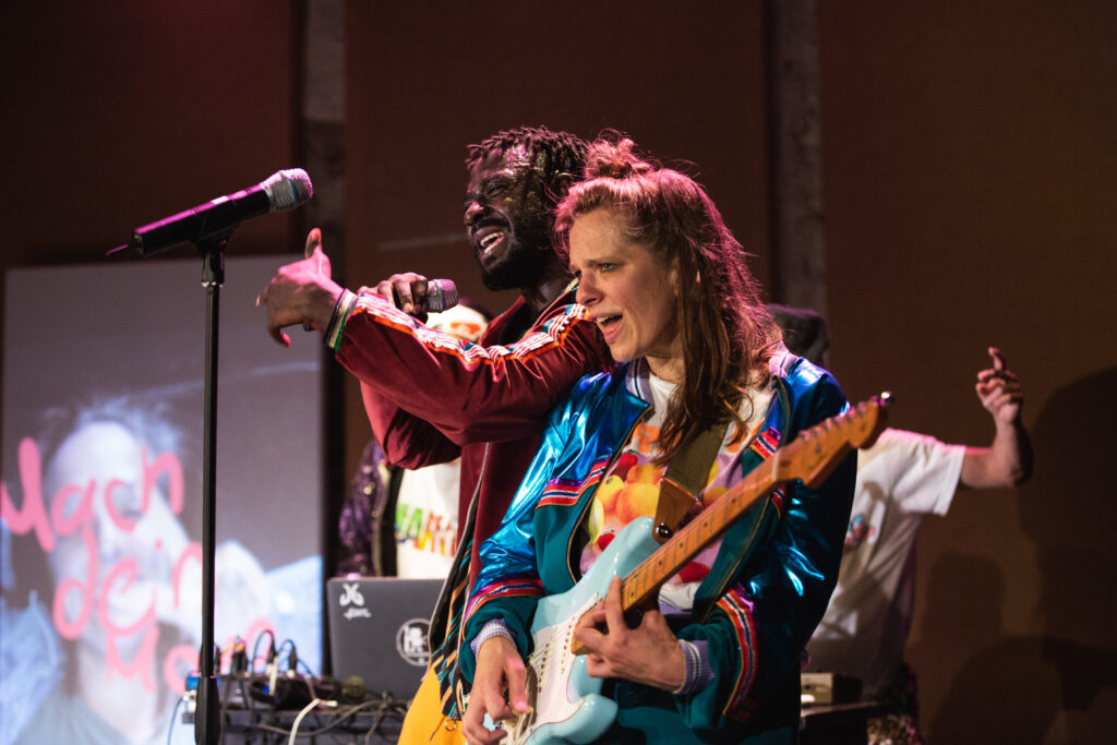 Kobieta gra na gitarze i śpiewa. Za nią śpiewający mężczyzna.