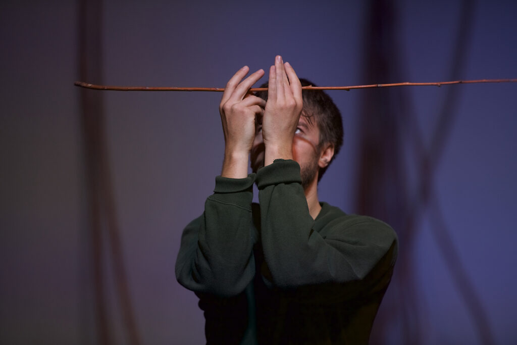 Mężczyzna trzyma na wysokości czoła jedną cienką witkę wierzbową. Ręce zasłaniają jego twarz.