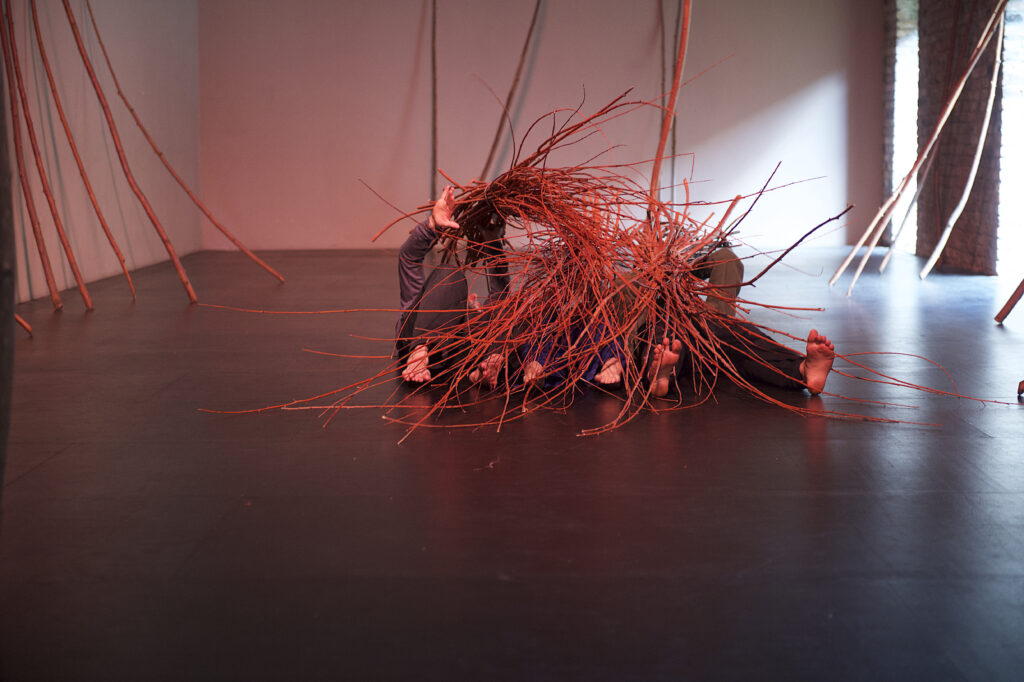 Trzy osoby leżą na podłodze przykryte witkami wierzbowymi jak kołdrą.