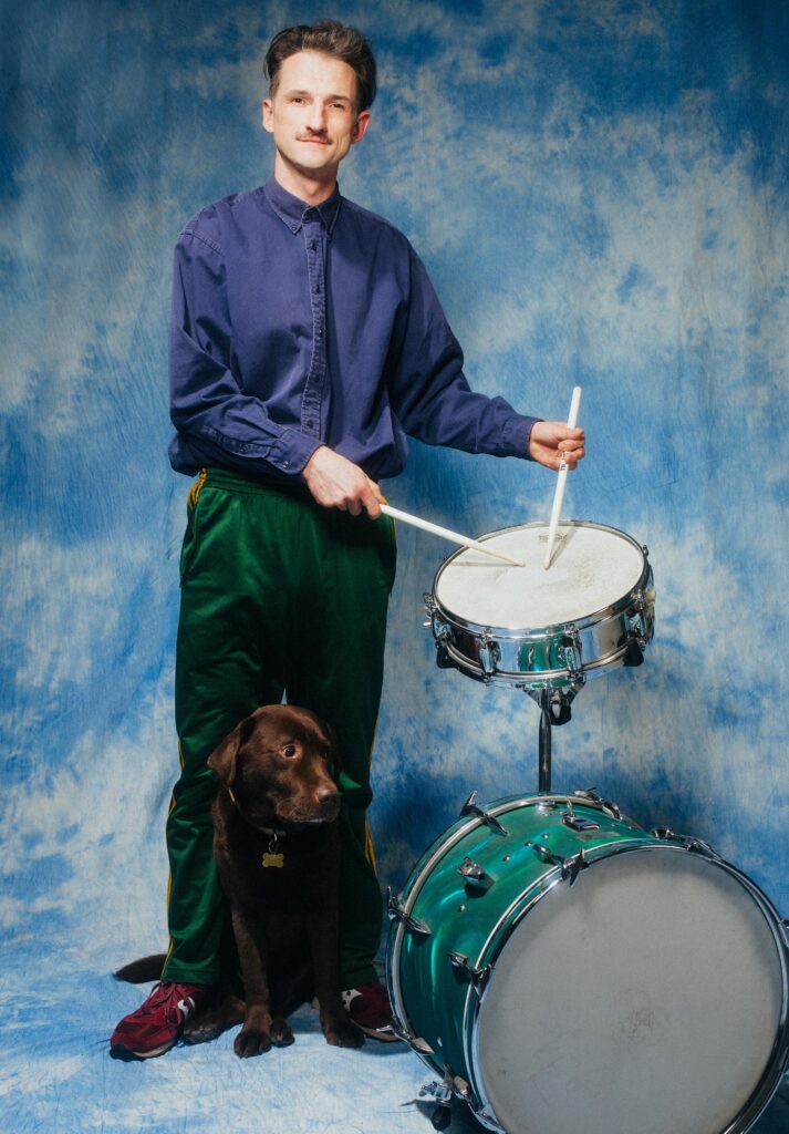 Mężczyzna pozuje do zdjęcia. Trzyma w rękach pałeczki i udaje, że gra na werblu. Między stopami mężczyzny siedzi uroczy labrador.