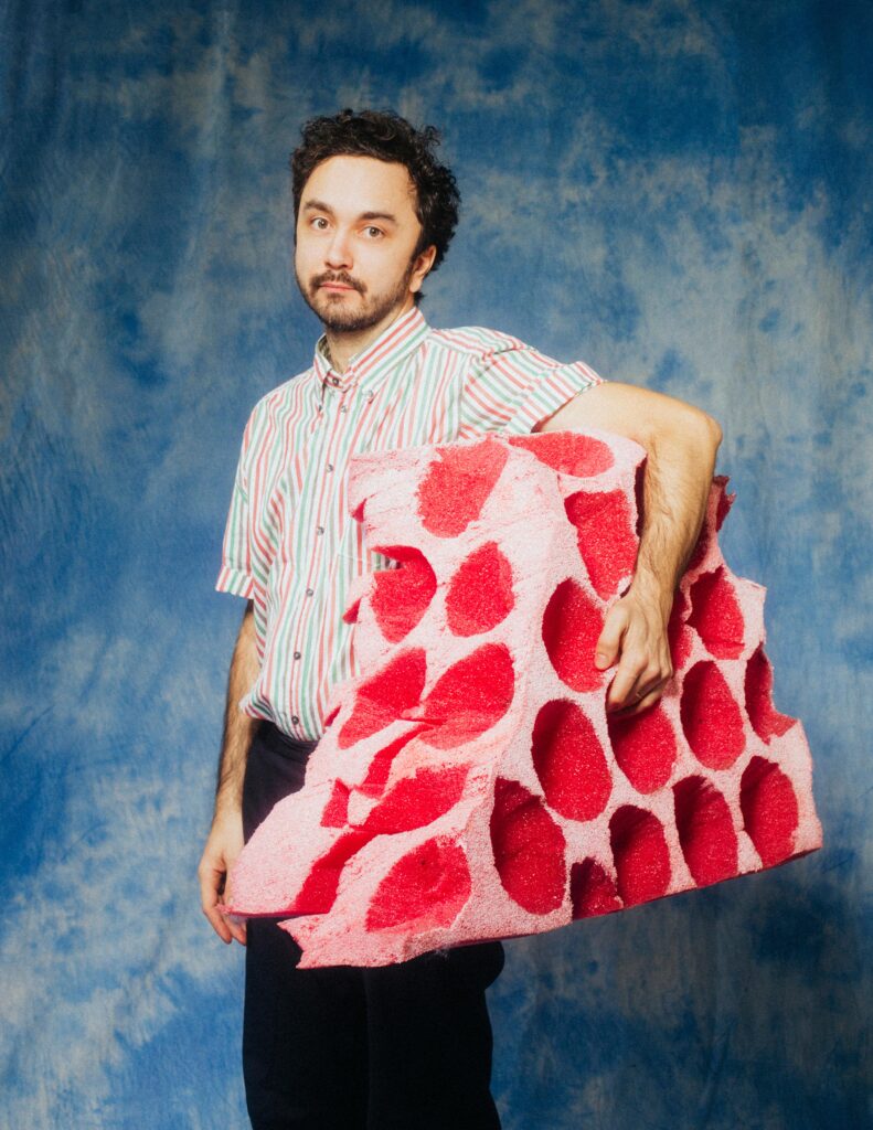 Mężczyzna pozuje od zdjęcia. Pod lewą ręką trzyma ogromną czerwoną gąbkę.