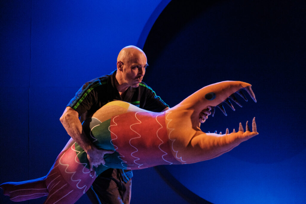 Mężczyzna trzyma w rękach wielką maskotkę przypominającą podłużną rybę. Ma ona wielkie zęby.