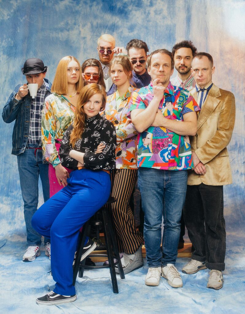 Dziesięć osób pozuje do zdjęcia. Z przodu kobieta siedzi na wysokim stołku. Po jej lewej stronie stoi mężczyzna w kolorowej koszuli i drapie się po brodzie. Z tyłu po prawej stronie stoi mężczyzna w czapce i pije herbatę. Trzy osoby po środku z tyłu mają okulary przeciwsłoneczne.