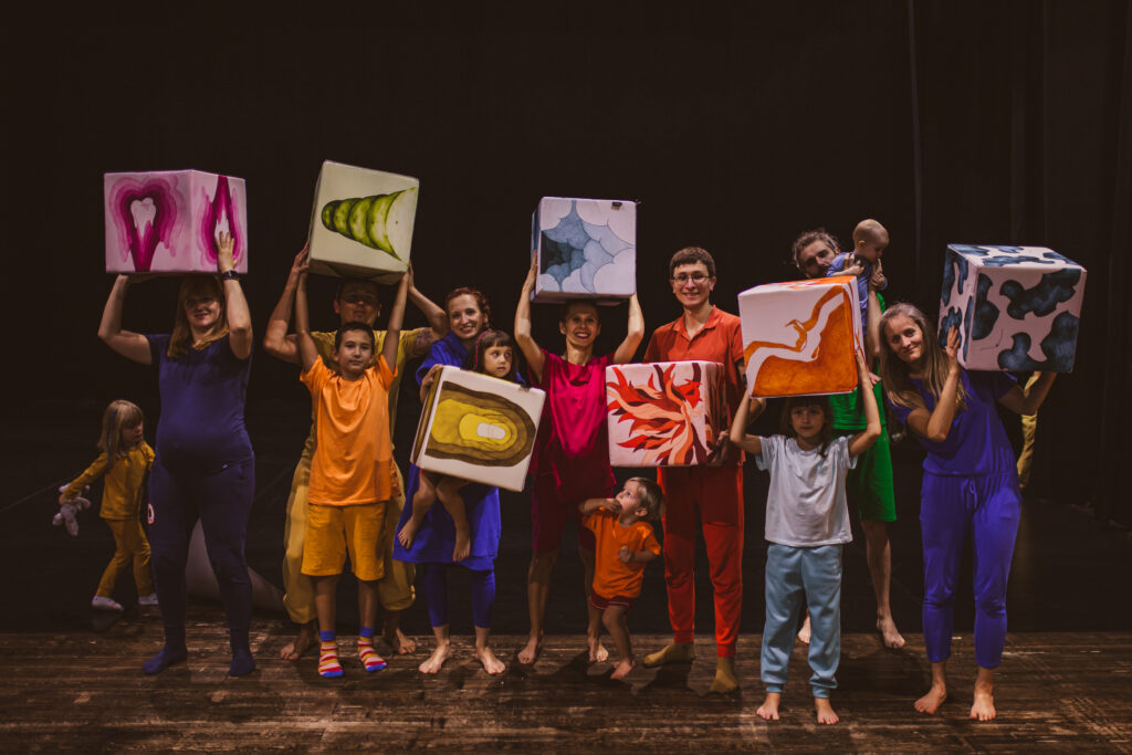 Ania Wańtuch i Performujące Rodziny. Do zdjęcia pozuje siódemka dorosłych i szóstka dzieci. Niektórzy trzymają w rękach wielkie kostki z kolorowymi rysunkami. Grupa jest kolorowa i uśmiechnięta.