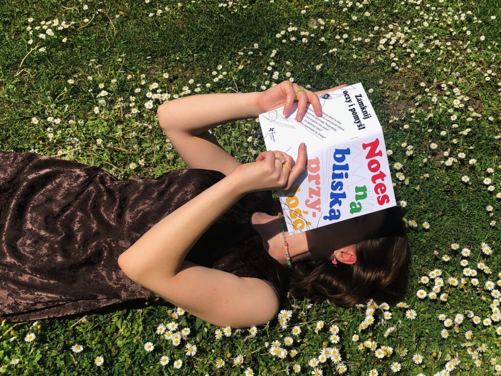 Kobieta leży na trawie, w rękach trzyma zeszyt z napisem: Notes na bliską przyszłość.