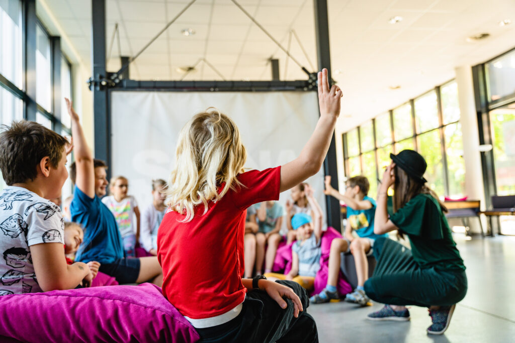 W budynku Sceny Wspólnej grupa dzieci siedzi. Przed nimi kuca kobieta w kapeluszu. Z przodu jedno z dzieci ma uniesioną rękę.
