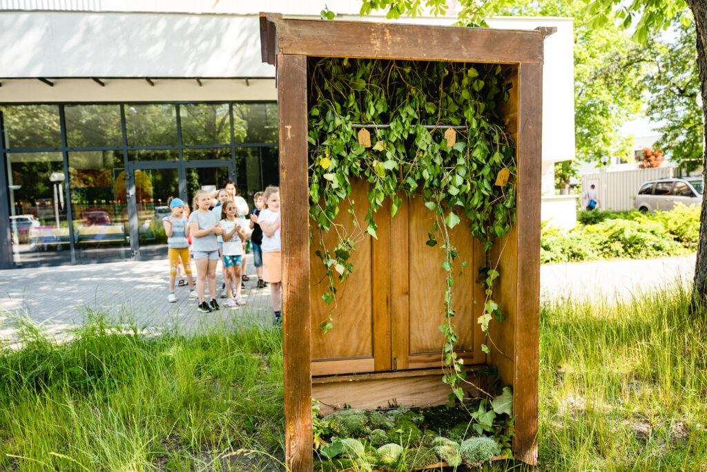 Z przodu szafa obwieszona liśćmi. Za nią w tle grupa dzieci.