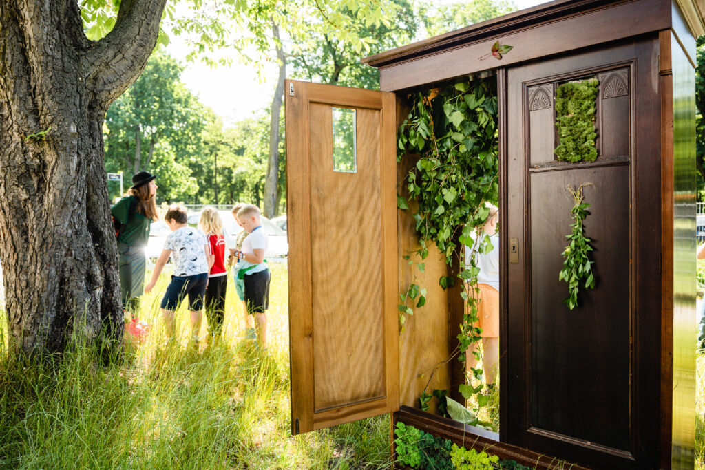 Po prawej otwarta szafa, obwieszone gałęziami i mchem. W tle kobieta w kapeluszu i grupa dzieci.