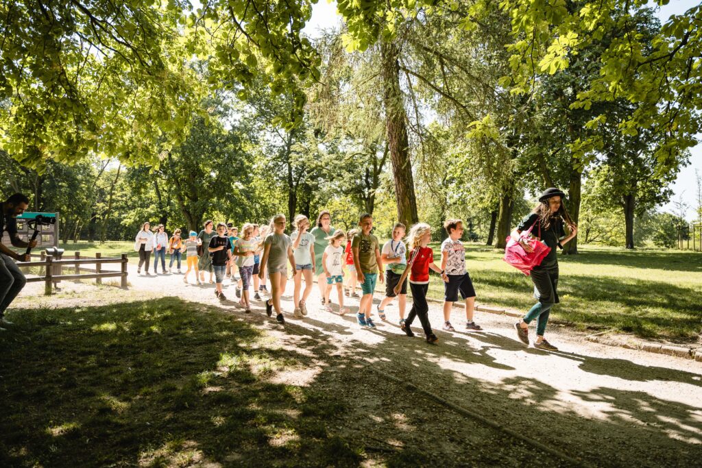 W lesie grupa dzieci spaceruje. Na przedzie kobieta w kapeluszu.