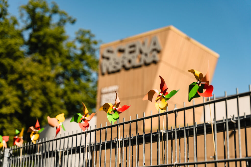 Płot, do którego przyczepione są wiatraczki. W tle budynek z napisem: Scena Wspólna. / A fence with small windmills attached to it. In the background a building with a sign: Scena Wspólna.