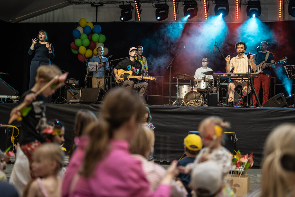 W tle na scenie grupa muzyków. Z przodu widownia.