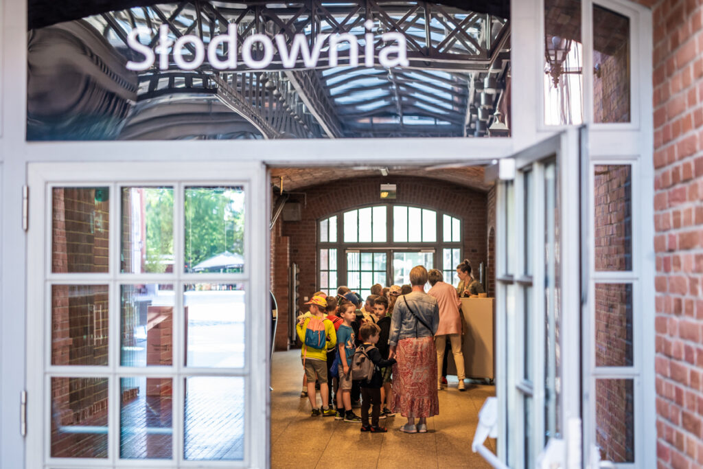 W tle grupa dzieci i dorosłych. Na przedzie otwarte drzwi. Nad nimi napis: Słodownia.