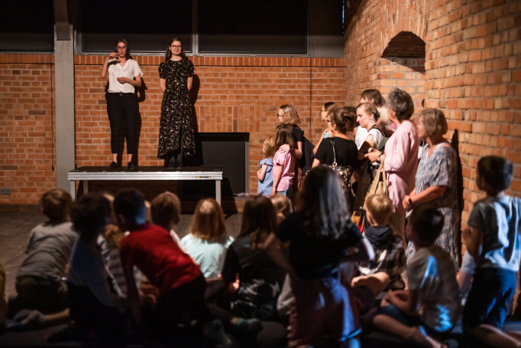 Grupa osób w sali. W tle po lewej dwie kobiety stoją na podwyższeniu. Jedna używa języka migowego.