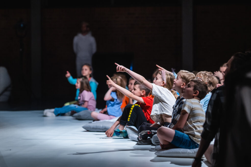 Grupa dzieci siedzi na podłodze i wskazuje palcami w lewo.