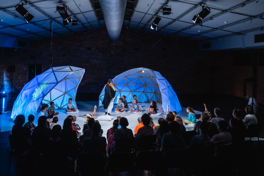 Na przedzie widownia. W tle scena, na której ustawiona dwie połówki igloo. Na środku aktorka ubrana w strój przypominający pingwina.