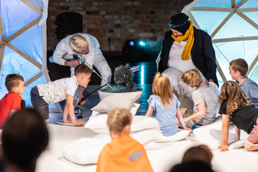 Na scenie grupa dzieci siedzi na poduszkach. Aktorka w stroju pingwina spogląda na kukiełkę ptaka.